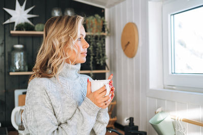 Young woman in grey sweater with cup of hot drink in hands looking at window on kitchen at house