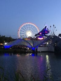 Illuminated ferris wheel by river against sky at night