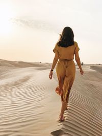 Rear view of woman walking on sand dune at desert against sky during sunset