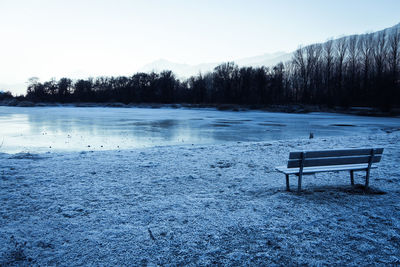 Empty bench by frozen lake against sky during winter