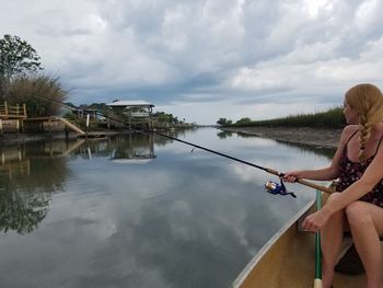 Young woman fishing while traveling in boat on river against cloudy sky