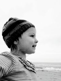 Thoughtful girl wearing knit hat while smiling at beach against clear sky