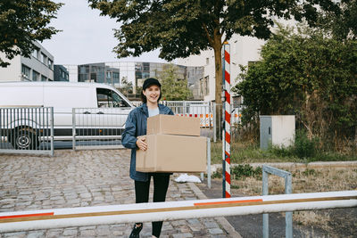 Smiling female delivery person holding cardboard boxes while walking on footpath