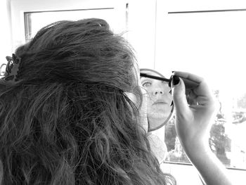 Rear view of woman using mirror while applying mascara at home