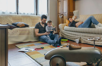 Mother reading book while father using digital tablet with son in living room