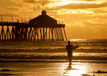 Silhouette woman holding surfboard on shore against cloudy sky during sunset