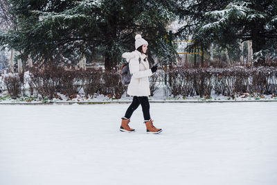 Caucasian young woman walking outdoors in city during winter while snowing, using mobile phone