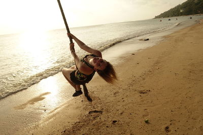 Full length portrait of smiling young woman hanging on rope at beach against sky during sunset