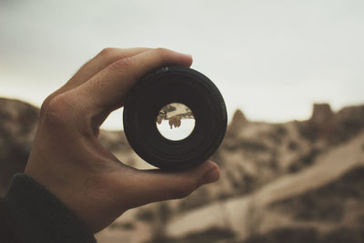 Cropped image of hand holding camera lens in front of mountain