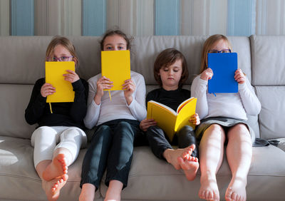 Four children sitting comfortably together on sofa in living room and doing prep work for school