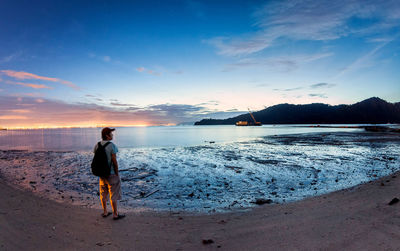 Rear view of man standing at beach against sky during sunset