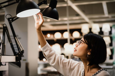 Asian young beautiful woman wearing glasses choosing new lamp to buy at furnishings lighting store