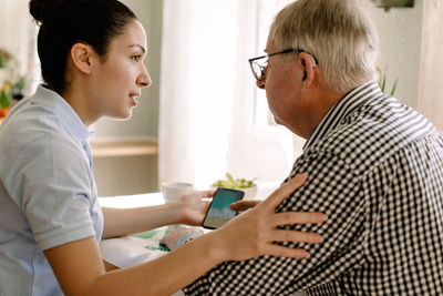 Young female social worker assisting retired man with smart phone in nursing home