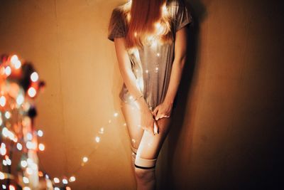 Midsection of woman standing with illuminated string lights against wall