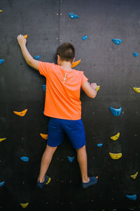 Back view of unrecognizable fearless child balancing on grips and climbing wall while practicing bouldering in gym