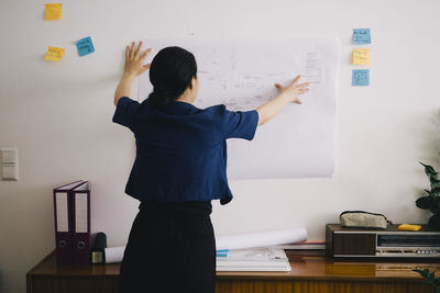 Rear view of female architect examining blueprint on wall at home office