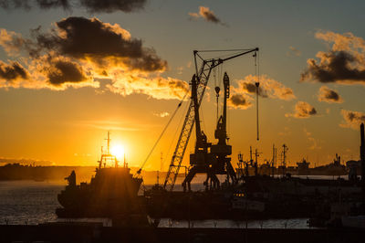 Silhouette cranes at harbor against sky during sunset