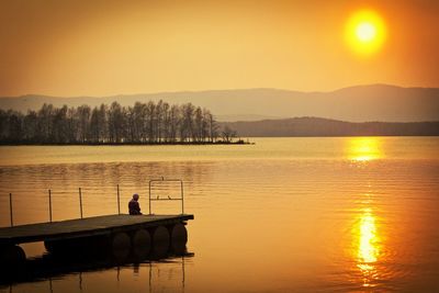 Person sitting on pier over river during sunset