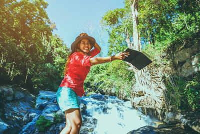 Smiling young woman holding book while standing by waterfall