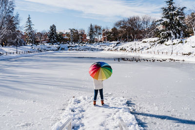 Back view of woman holding colorful umbrella standing on snowy pier by frozen lake during winter