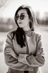 Confident young woman with arms crossed wearing sunglasses and jacket while standing outdoors