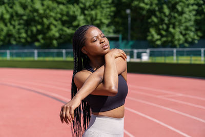 Black young woman with long braids stretches hands and shoulders standing on red track on stadium