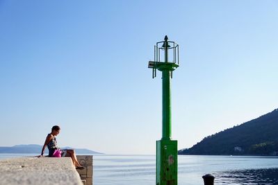 Side view of woman sitting on pier by sea against clear sky