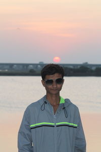 Portrait of man wearing sunglasses standing against sea during sunset