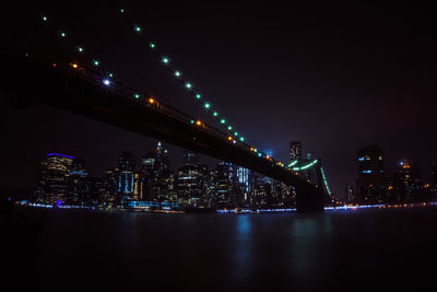 Illuminated bridge over river by buildings against sky at night