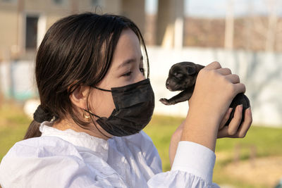 A girl in a protective mask on her face is holding a newborn labrador puppy. 