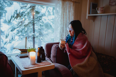 Side view of young woman in eyeglasses under coverlet resting on sofa with mug near window with view of fir woods in snow
