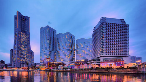 Modern buildings by river against sky in city at dusk