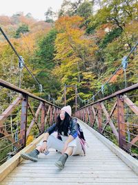 Full length of woman sitting on footbridge against trees at forest during autumn