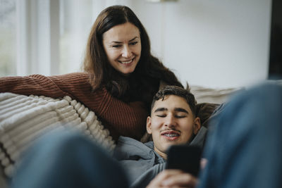 Smiling boy sharing smart phone with mother while lying on sofa in living room at home