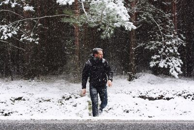 Man crossing while looking away during snowfall against trees in forest