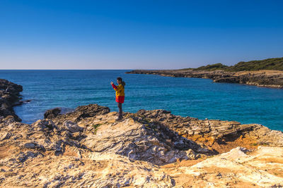 Side view of man standing on rock formation by sea against clear blue sky