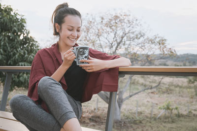 Portrait of young woman sitting on bench against sky