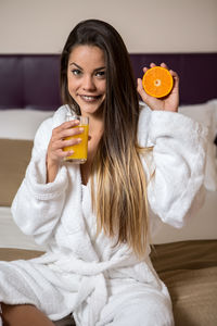 Portrait of smiling beautiful woman holding drink and fruit while sitting on bed at hotel