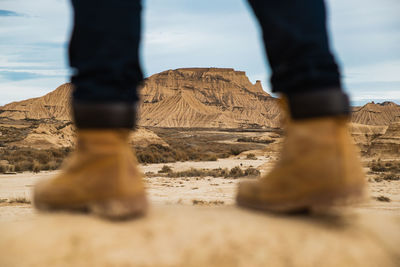 Legs on faceless traveler in brown boots and blue jeans standing on dirty sandy road with mountain and sky on blurred background in bardenas reales, navarre, spain