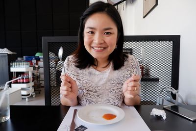 Portrait of smiling young woman holding cutleries eating food while sitting in restaurant