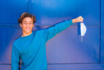 Smiling teenage boy holding protective mask against wall