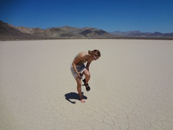 Full length of young woman taking sandals off at racetrack playa against sky, death valley