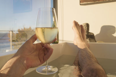 Low section of man holding wine glass while sitting in bathtub