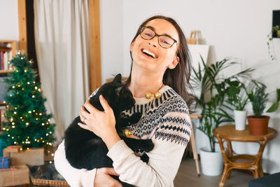 Young woman in cozy sweater hugging black domestic cat and smiling in the room with christmas tree.