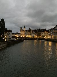 Illuminated buildings by river against cloudy sky