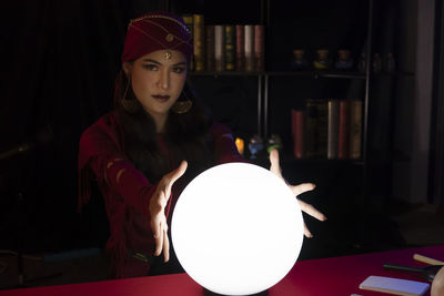 Young woman gesturing over crystal ball while sitting in darkroom