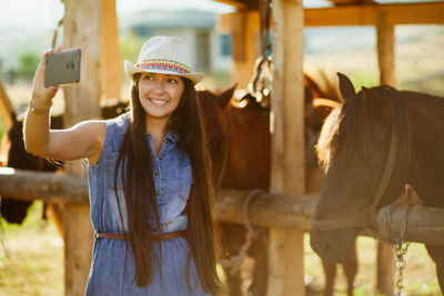 Smiling young woman taking selfie by horses at pen