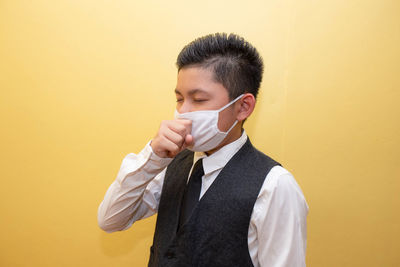 Boy wearing mask coughing while standing against yellow wall
