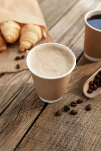 Two paper cups of coffee, croissants bundle at sackcloth, dried roasted coffee beans at wooden table