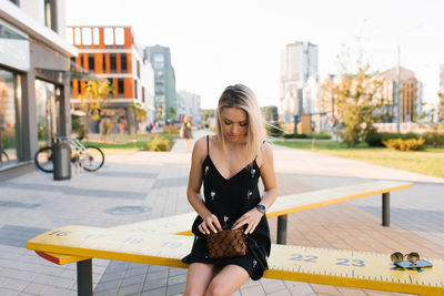Blonde woman in a black dress is sitting on a bench in the city and is looking for something 
